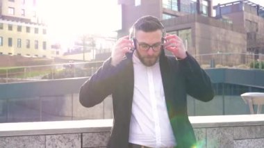 Beyaz sakallı genç adam dışarıda müzik dinliyor kulaklıklarla ve akıllı telefon ahizesiyle mesajlaşıyor gülümsüyor ve rahatlıyor.