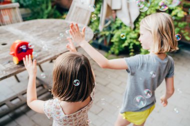 Bayan çocuklar evlerinin terasında sabun köpüğüyle oynuyorlar. Birlikte vakit geçiriyorlar.