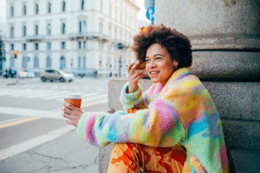 Geleneksel olmayan farklı etnik çeşitlilikte genç bir kadın dışarıda oturmuş kahve içiyor ve anlık sesli mesajlar dinliyor.