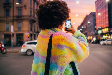 Açık havada akıllı telefon kullanan çok ırklı genç bir kadın şehirde günbatımının resmini çekiyor.