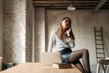 Young asatic professiona businesswoman sitting on table indoor at home using computer and talking smartphone