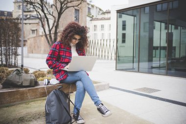 Genç melez kıvırcık saçlı kız kampüste bilgisayar kullanarak ders çalışıyor.