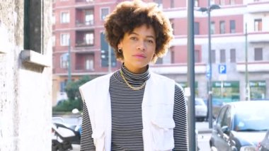 Genç, kıvırcık saçlı, dışarıda gezen, kameraya sağlıklı gülümseyen, özgür ve kendinden emin siyah bir kadının ağır çekimde portresi.