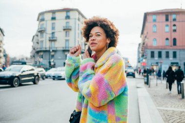 Genç kıvırcık saçlı, açık havada konuşan, akıllı telefonlu, eğlenen bir kadın.