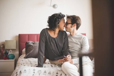 Genç, yakışıklı melez bir çift yataklarında oturuyor, akıllı telefon kulpu kullanıyor, göz göze bakıyor, gülümsüyor - teknoloji, sosyal ağ, iletişim konsepti