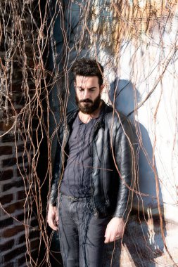 Sakallı genç adam dışarıda poz veriyor.