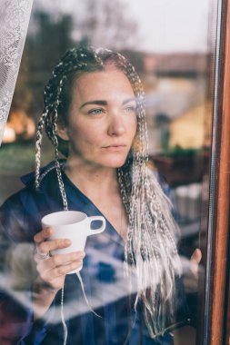 Pencereden dışarı bakan yetişkin bir kadın elinde pijamalarıyla geleceğe bakıyor.