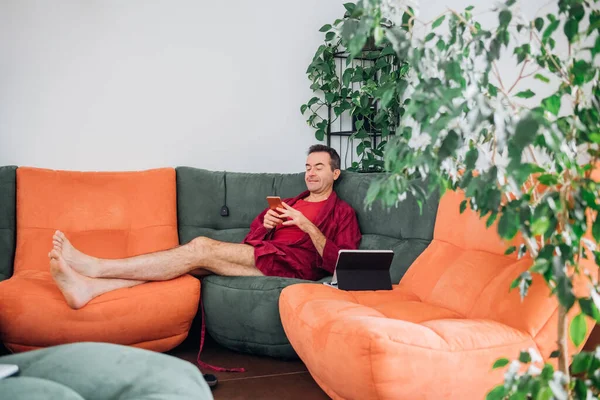 Single man indoor at home relaxing on sofa, using smartphone and tablet