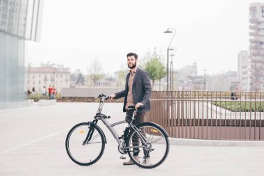 Çağdaş sakallı genç işadamı, elinde bisikletle dışarıda poz veriyor, gülümsüyor.