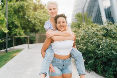 İki genç, etnik çeşitliliğe sahip lezbiyen çift kucaklaşıyorlar, sırtına binip gülüyorlar.