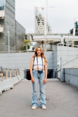 Kameralı beyaz kadın dışarıda poz veriyor. Ciddi bir tavır sergiliyor.