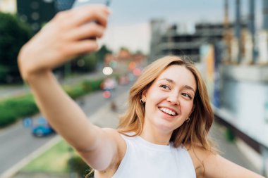 Genç beyaz kadın dışarıda akıllı telefon kullanıyor. Mutlu bir şekilde selfie çekiyor.