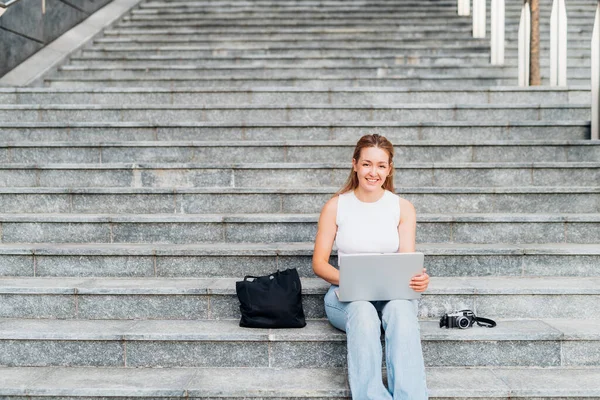 Young caucasian woman digital nomad remote working outdoor sitting staircase using computer smiling happy and serene