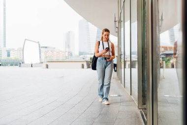 Beyaz kadın turist dışarı çıkıp gülümsüyor ve akıllı telefon kullanarak vitrinlere bakıyor.