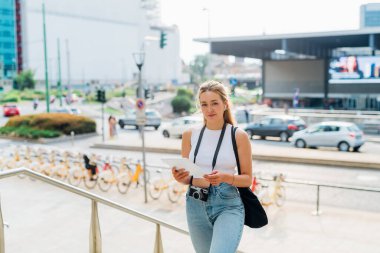 Genç beyaz kadın kulaklık ve tablet kullanarak dışarıda yürüyor sesli kitaptan zevk alıyor ya da serbest olarak çalışıyor.