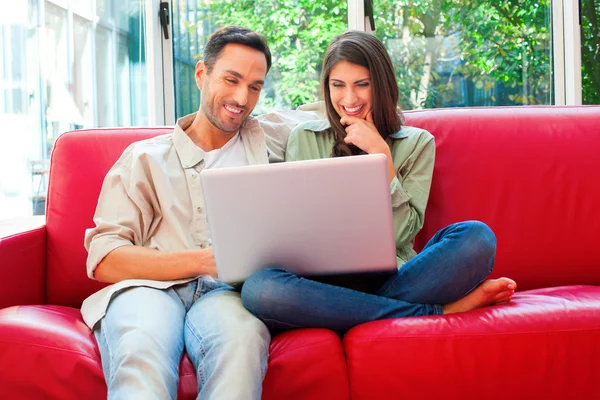 Happy young couple using laptop on red sofa — Stock Photo, Image