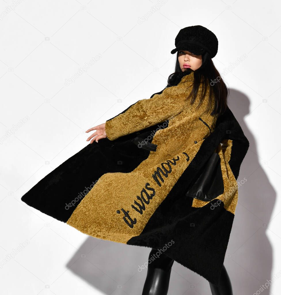 Stylish woman in two-tone karakul fur coat, leather pants and fur cap stands back to camera, spinning