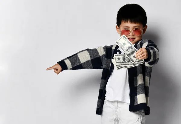 Asiático niño en gafas de sol, cárdigan a cuadros y pantalones blancos muestra manojo fan de dólares dinero en efectivo y puntos a un lado —  Fotos de Stock