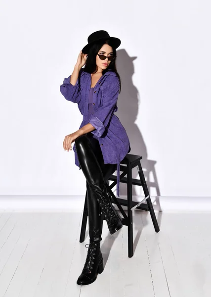 Hermosa mujer en pantalones de cuero negro, chaqueta de camisa púrpura, sombrero negro y gafas de sol se sienta en la silla y mira a un lado —  Fotos de Stock