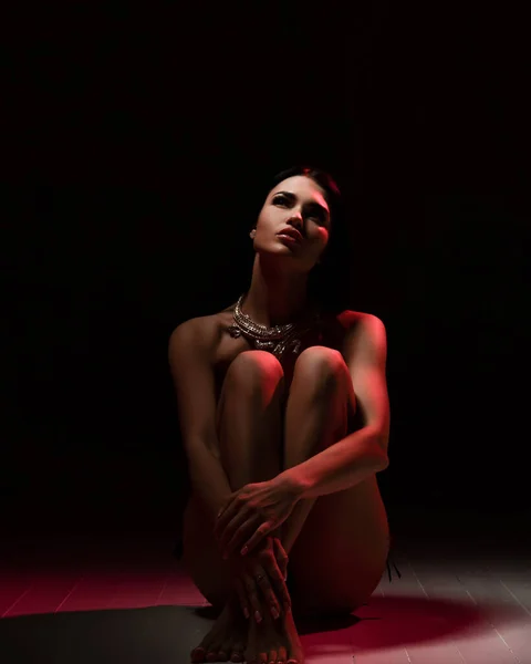 Sensual mujer romántica desnuda en collar de joyería se sienta en el suelo con los brazos alrededor de sus rodillas y mira hacia arriba — Foto de Stock