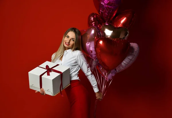 Mooie blonde vrouw met cadeau doos en paars roze hart ballonnen voor nieuwjaar viering gelukkig glimlachen op donker rood — Stockfoto