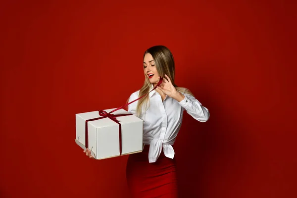 Emocionado mujer rubia feliz en blusa blanca desempaca gran caja de regalo para San Valentín Día sobre fondo rojo oscuro — Foto de Stock