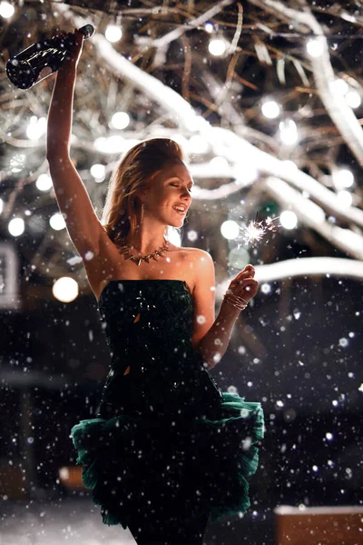 Hermosa chica de pelo rojo con bengalas encendidas y una botella de champán celebra alegremente la Navidad y el año nuevo por la noche — Foto de Stock