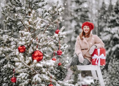 Süslü, sıcak giysiler içinde gülümseyen genç bir kadın Noel hediyesiyle dışarıda, Noel 'in yanında, taburede oturuyor.