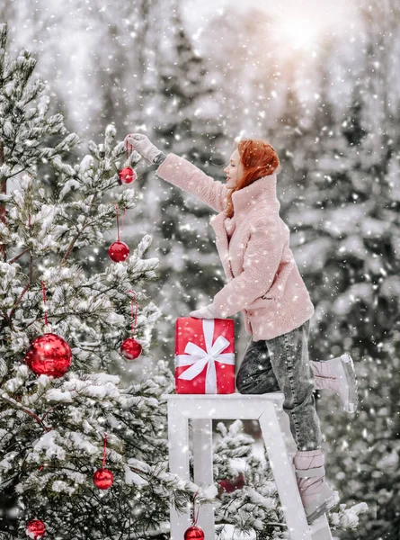 Young woman in warm clothing pink fur coat standing and decorating Christmas tree with balls outdoors in winter snowy forest