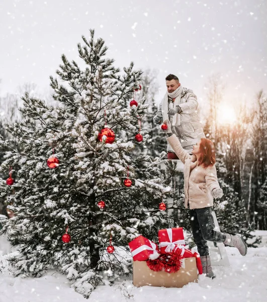 Junges glückliches Paar in stylischer Winterkleidung schmückt Weihnachtsbaum mit roten Kugeln im winterlich verschneiten Wald — Stockfoto