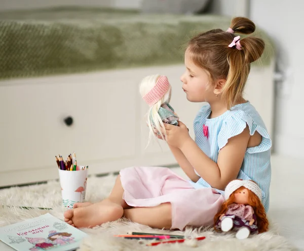 Милая маленькая девочка в домашней одежде сидит, играет с куклами и рисует карандашами дома — стоковое фото