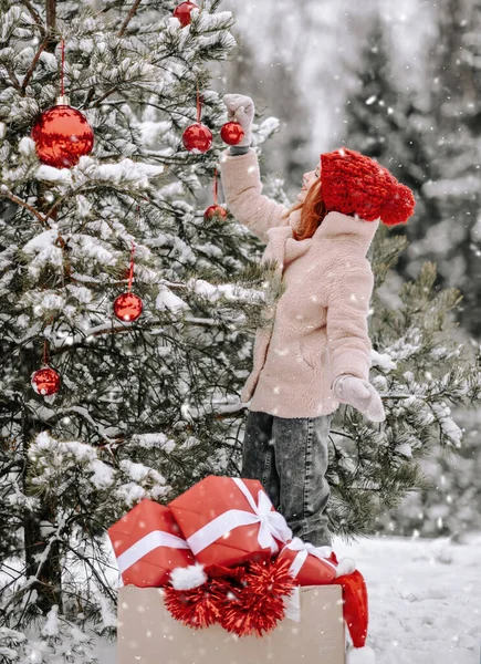 Mladá šťastná hezká žena ve stylové zimní oblečení zdobí venkovní vánoční strom jedle s červenými kuličkami — Stock fotografie