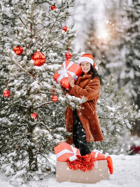 화려 한 모피 코트를 입은 젊은 섹시 한 갈색 여성이 크리스마스 트리에 서서 붉은 선물 상자를 껴안고 있다. — 스톡 사진