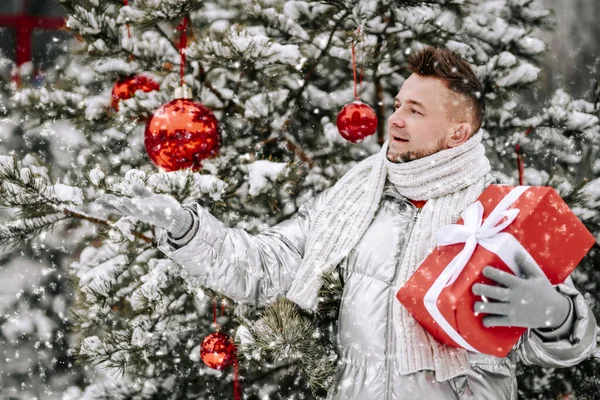 행복 한 턱수염을 가진 남자가 크리스마스 트리에 서 붉은 보철 상자를 들고 야외에서 눈송이를 잡고 — 스톡 사진
