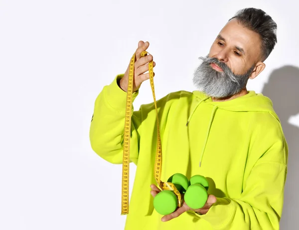 Mature hipster old brutal bearded man in yellow hoodie hold green dumbbells and tape measure with white stylish beard on white — Stock Photo, Image