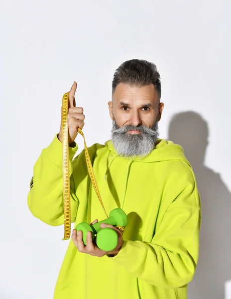 Mature hipster old brutal bearded man in yellow hoodie hold green dumbbells and tape measure with white stylish beard on white — Stock Photo, Image