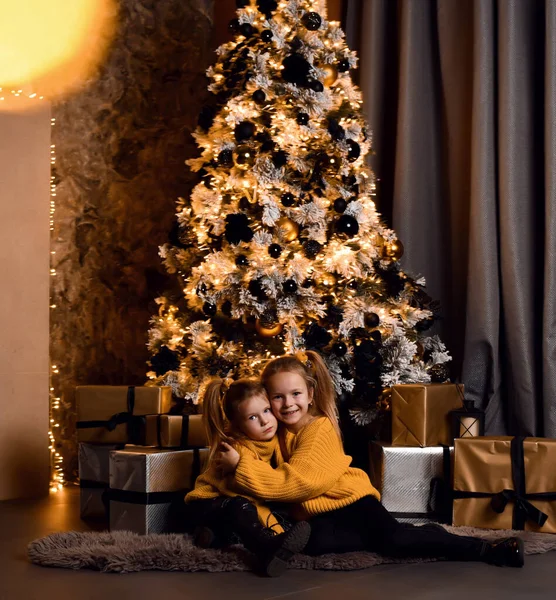 Gelukkig kleine meisjes zussen in geel truien zitten op de vloer samen, knuffelen kijken naar camera over versierde kerstboom Rechtenvrije Stockafbeeldingen