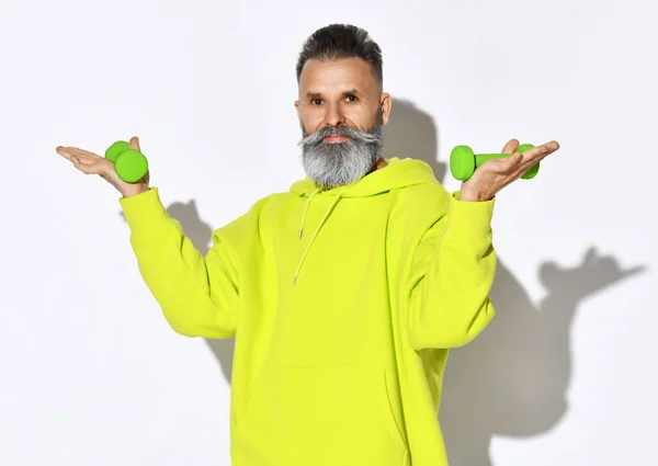 Portrait of happy active old brutal bearded man in yellow hoodie holding green dumbbells in hands on open palms — Stock Photo, Image
