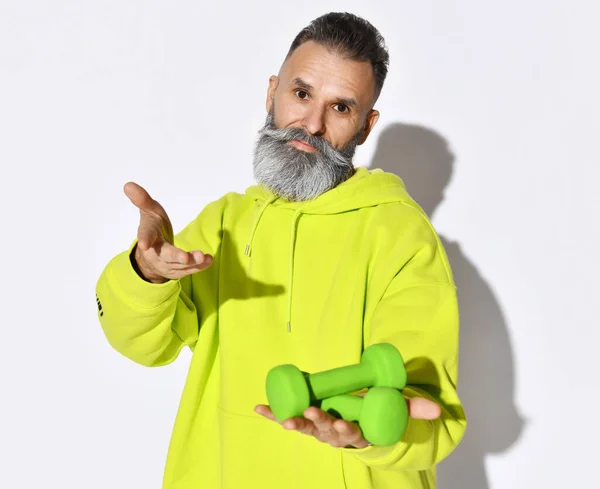 Active old brutal bearded man in yellow hoodie holding two green dumbbells in hand on open palm and calls to join — Stock Photo, Image