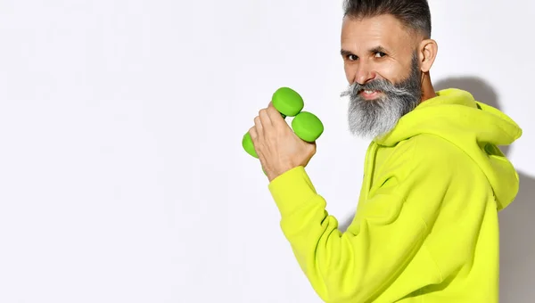 Portrait of cheerful active old brutal bearded man in yellow hoodie holding two green dumbbells in hand. Side view — Stock Photo, Image