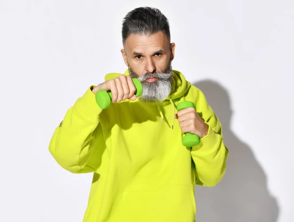 Sarı kapüşonlu yaşlı, vahşi sakallı adamın portresi. Elinde yeşil dambıllarla fitness antrenmanı yapıyor. — Stok fotoğraf
