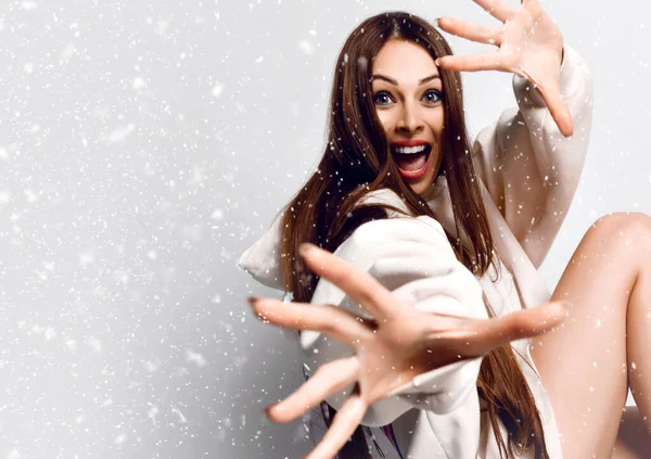 Alegre mujer excitada con el pelo largo en sudadera con capucha se sienta gesto con las manos y mirando a la cámara debajo de la nieve — Foto de Stock