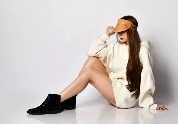 Sexig rik ung kvinna med långt hår i överdimensionerad huvtröja, stövlar sitter med nakna ben håller visir av sin lädermössa — Stockfoto