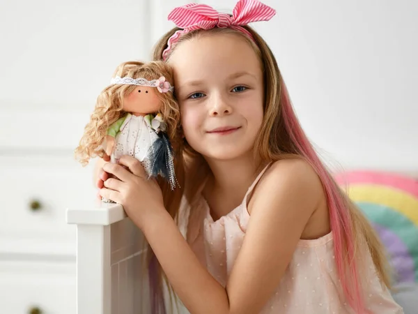 Счастливая девочка в домашней одежде и головной убор с луком играет фотографируется с ее любимой куклой дома — стоковое фото