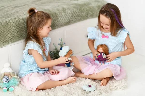 Две позитивные активные девочки в голубых платьях играют вместе со своими куклами на мягком ковре дома — стоковое фото