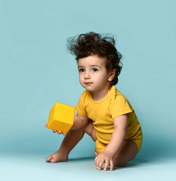 Маленький милий кучерявий волохатий дитина хлопчик малюк в жовтому зручному комбінезоні сидить на підлозі і грає з жовтою кубиковою іграшкою — стокове фото