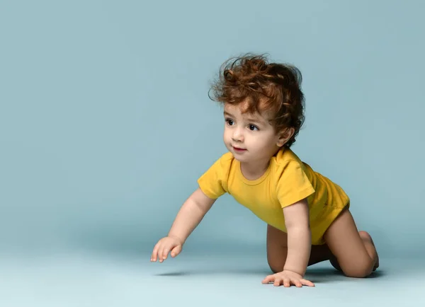 Şirin gülümseyen kıvırcık çocuk sarı rahat tulumuyla yerde sürünüyor ve neşeli hissediyor. — Stok fotoğraf