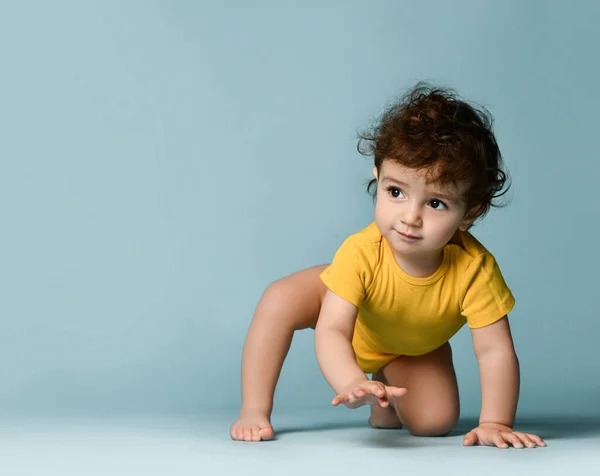 Küçük tatlı, pozitif kıvırcık çocuk sarı rahat tulumuyla yerde sürünüyor ve ayağa kalkmaya çalışıyor. — Stok fotoğraf
