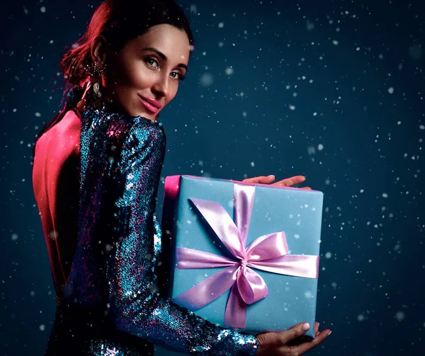 Young beautiful positive brunette woman in glittering black dress standing backwards with holiday present gift box in hands