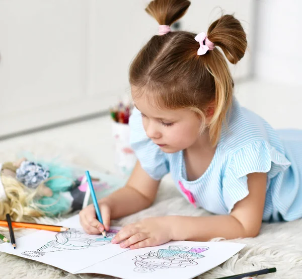 Menina criança concentrada calma está deitada no tapete macio desenho colorir fotos de bonecas com lápis coloridos — Fotografia de Stock